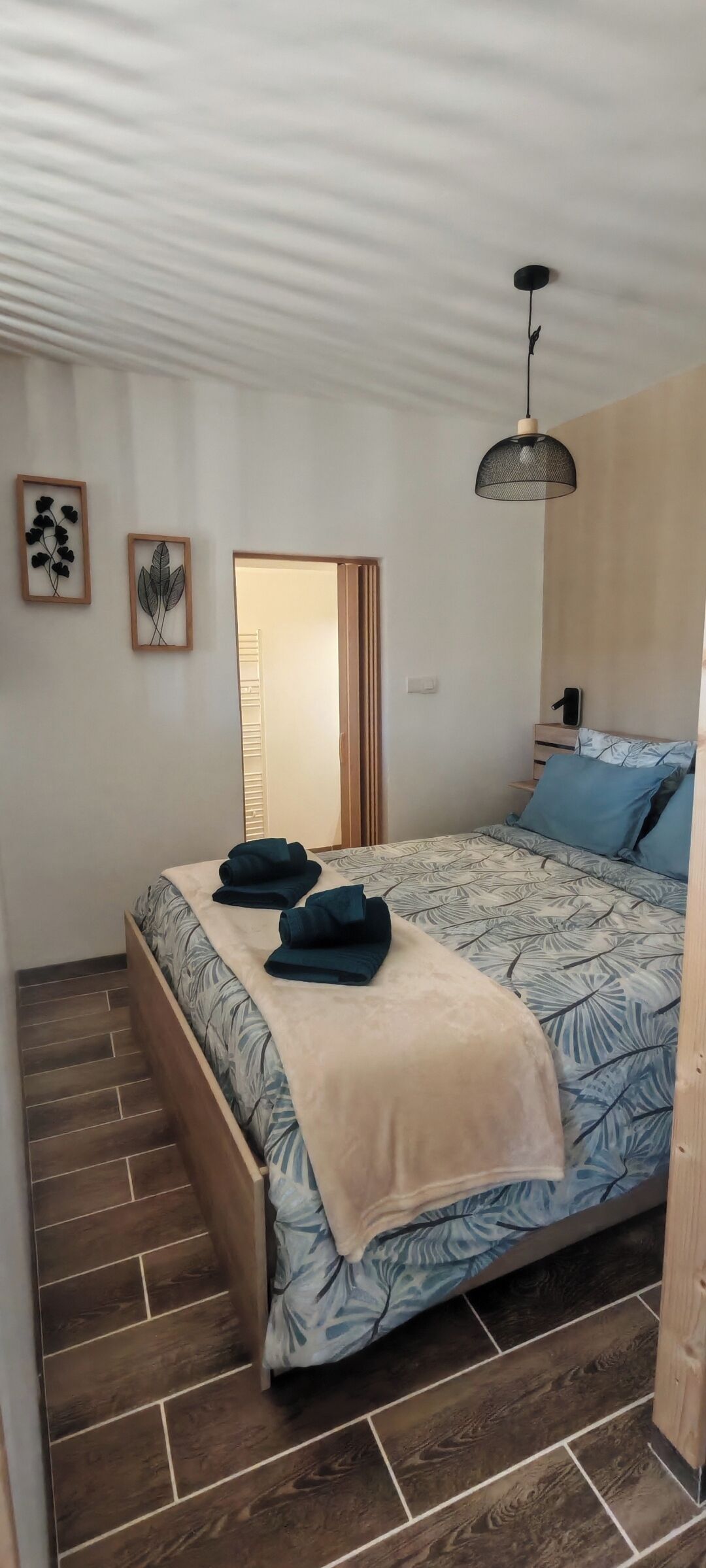A neatly arranged bedroom with a bed, a bedside table, a hanging light fixture, and a door leading to another room.