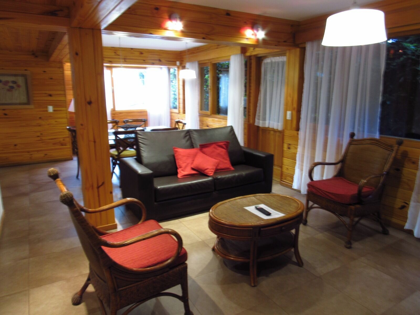 A cozy room with a wooden interior, featuring a black leather sofa with red cushions, two wooden chairs with red cushions, and a small round table.