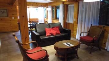A cozy room with a wooden interior, featuring a black leather sofa with red cushions, two wooden chairs with red cushions, and a small round table.