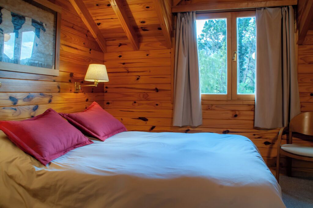 Une pièce de style cabane en bois, avec un lit, une lampe et une fenêtre avec des rideaux.