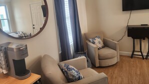 Ein Wohnzimmer mit Deckenventilator, einer Kaffeemaschine, zwei Sesseln, einem Fernseher und einem Klavier.