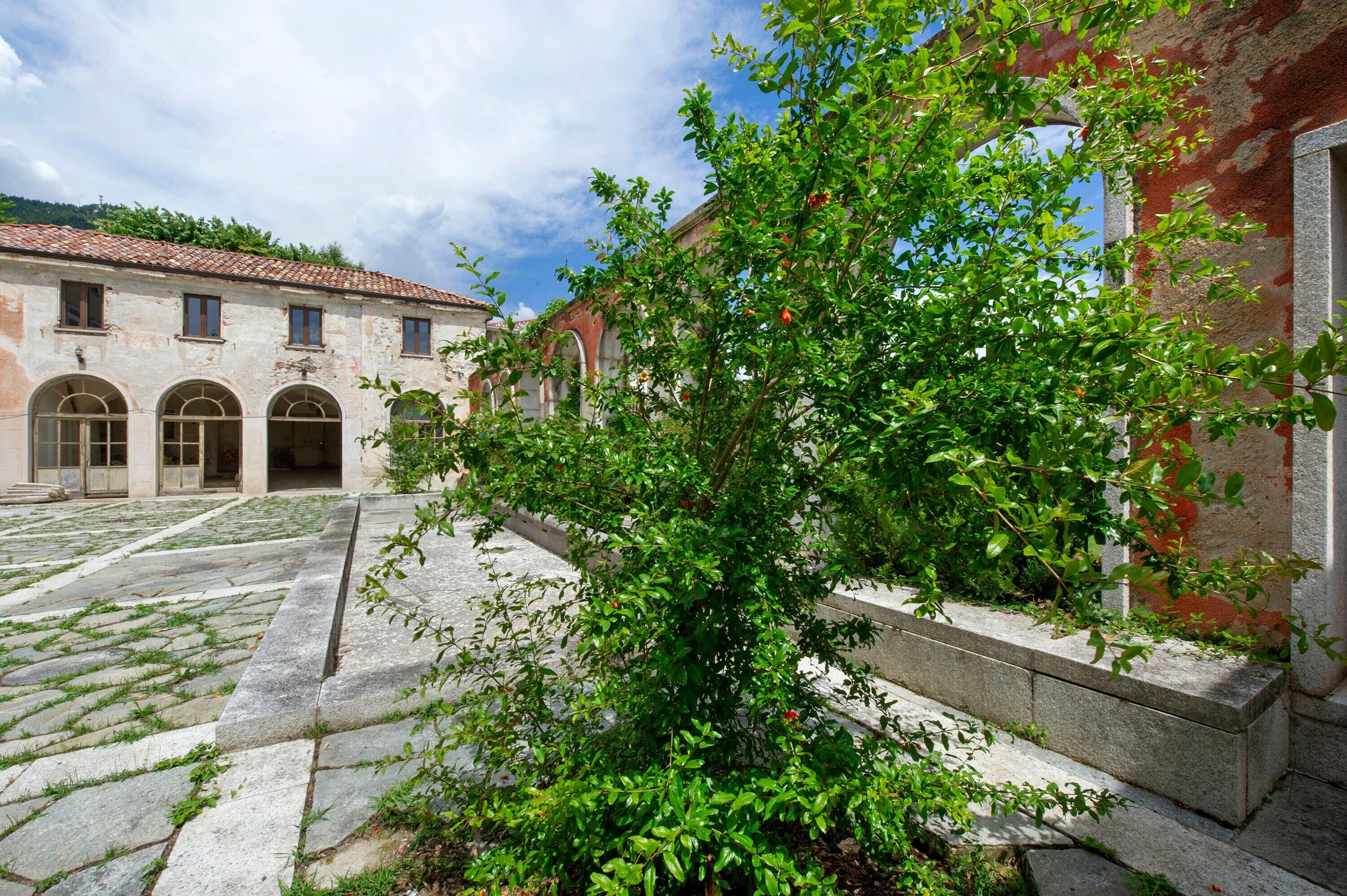 A courtyard with a building featuring arched doorways and a tiled roof, surrounded by greenery.