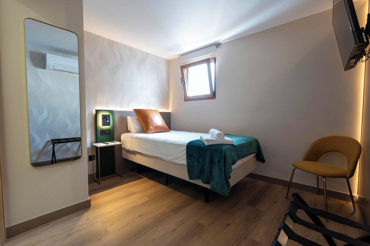 A bedroom with a bed, a chair, a bedside table, a mirror, and a window.