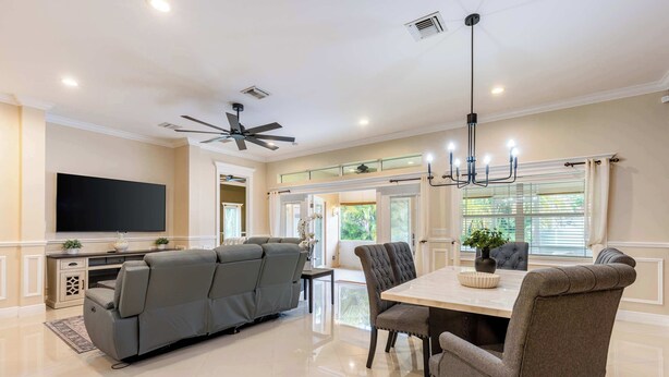 A modern living room with a sectional sofa, dining table, chairs, a flat-screen TV, ceiling fan, and large windows. - ‼️3BR Getaway Near Everything •Fenced Yard • BBQ (West Palm Beach)