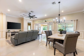 A modern living room with a sectional sofa, dining table, chairs, a flat-screen TV, ceiling fan, and large windows. - ‼️3BR Getaway Near Everything •Fenced Yard • BBQ (West Palm Beach)