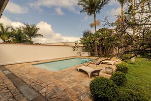 A swimming pool with stone tiles, surrounded by greenery and a white wall. - Ch Cara com Churrasqueira sal o de Jogos e Wi-fi (Suzano)