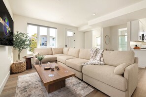 Una sala moderna con un sofá color beige, una mesa de centro de madera y un televisor montado en la pared.