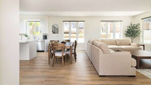 A spacious living area with a sofa, dining table, and chairs, a kitchen with white cabinets, and large windows.