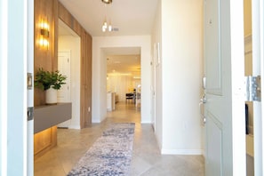 A hallway with a wooden accent wall, a potted plant, a blue and white rug, and a kitchen area in the background.
