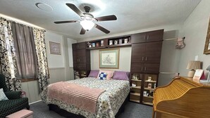 Un dormitorio con una cama, una mesita de noche de madera, un escritorio, un ventilador de techo y un armario.