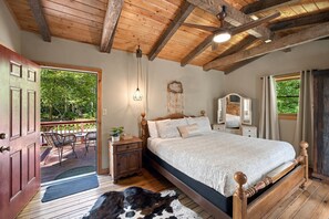 Un dormitorio con techo de madera, una cama con cabecera, una mesita de noche, un aparador, un espejo y vistas al exterior a través de una puerta.