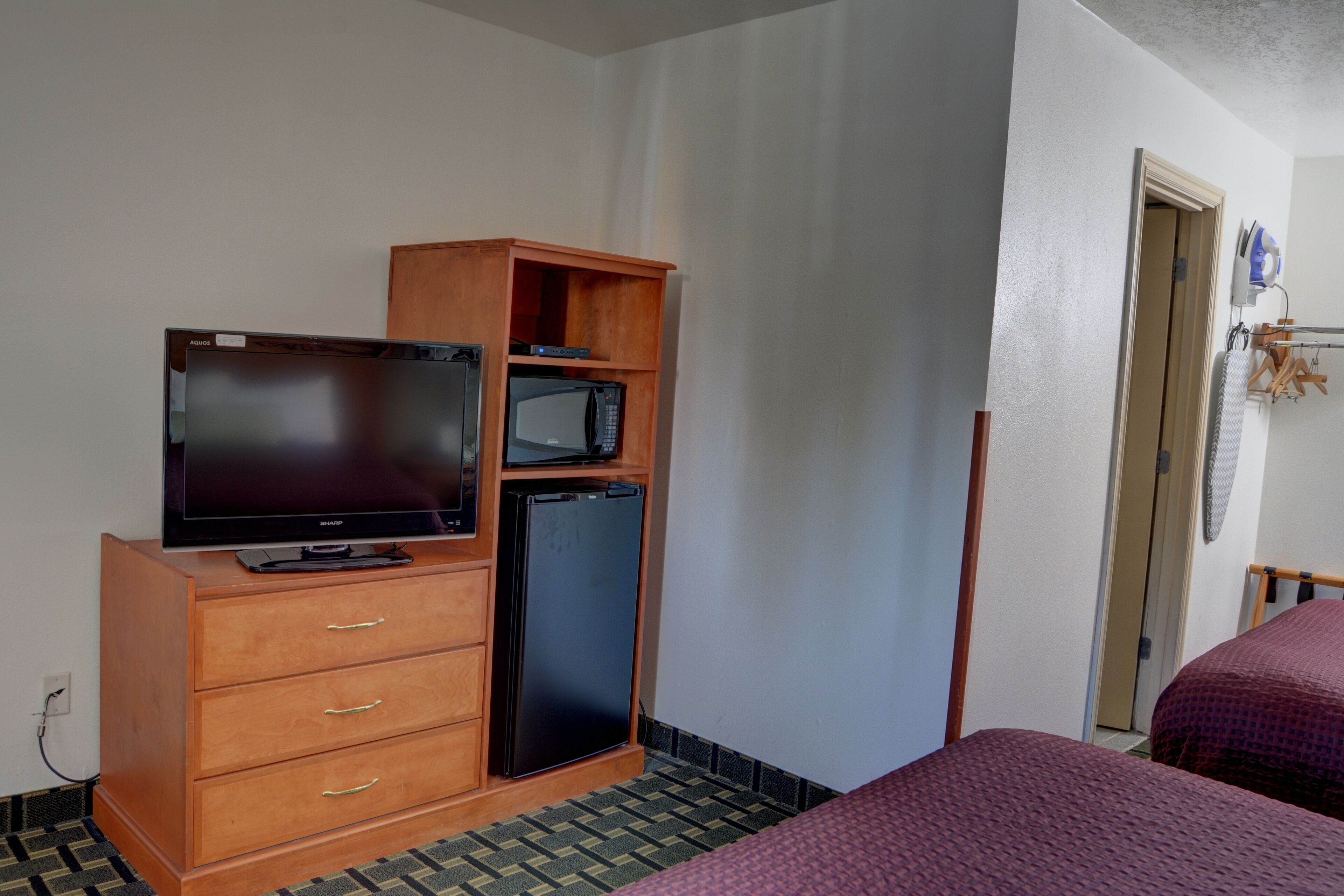 A hotel room with a wooden dresser, a television, a refrigerator, and two beds with purple bedding.