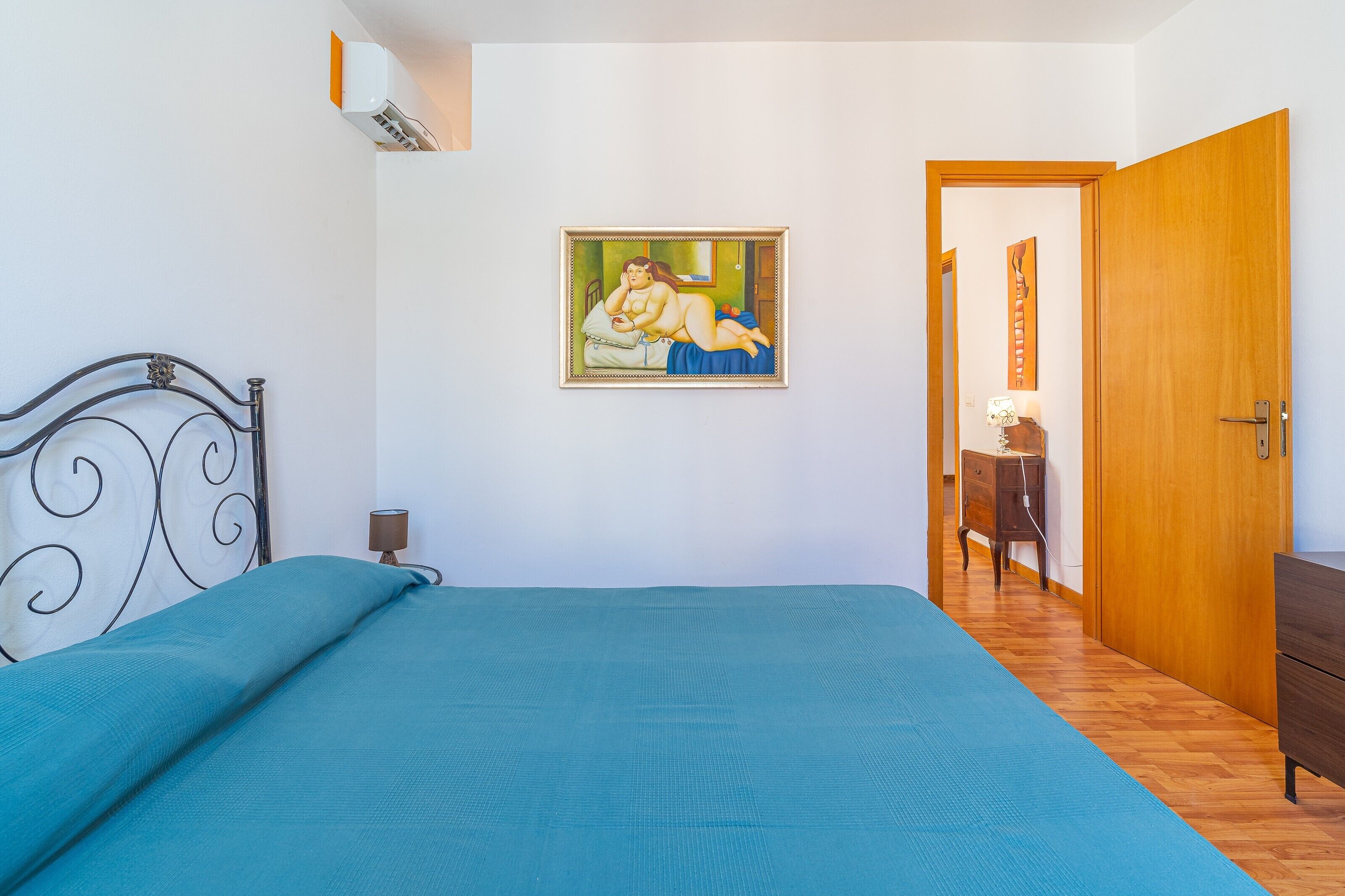 A bedroom with a bed, a painting, a wooden wardrobe, and a desk with a lamp.