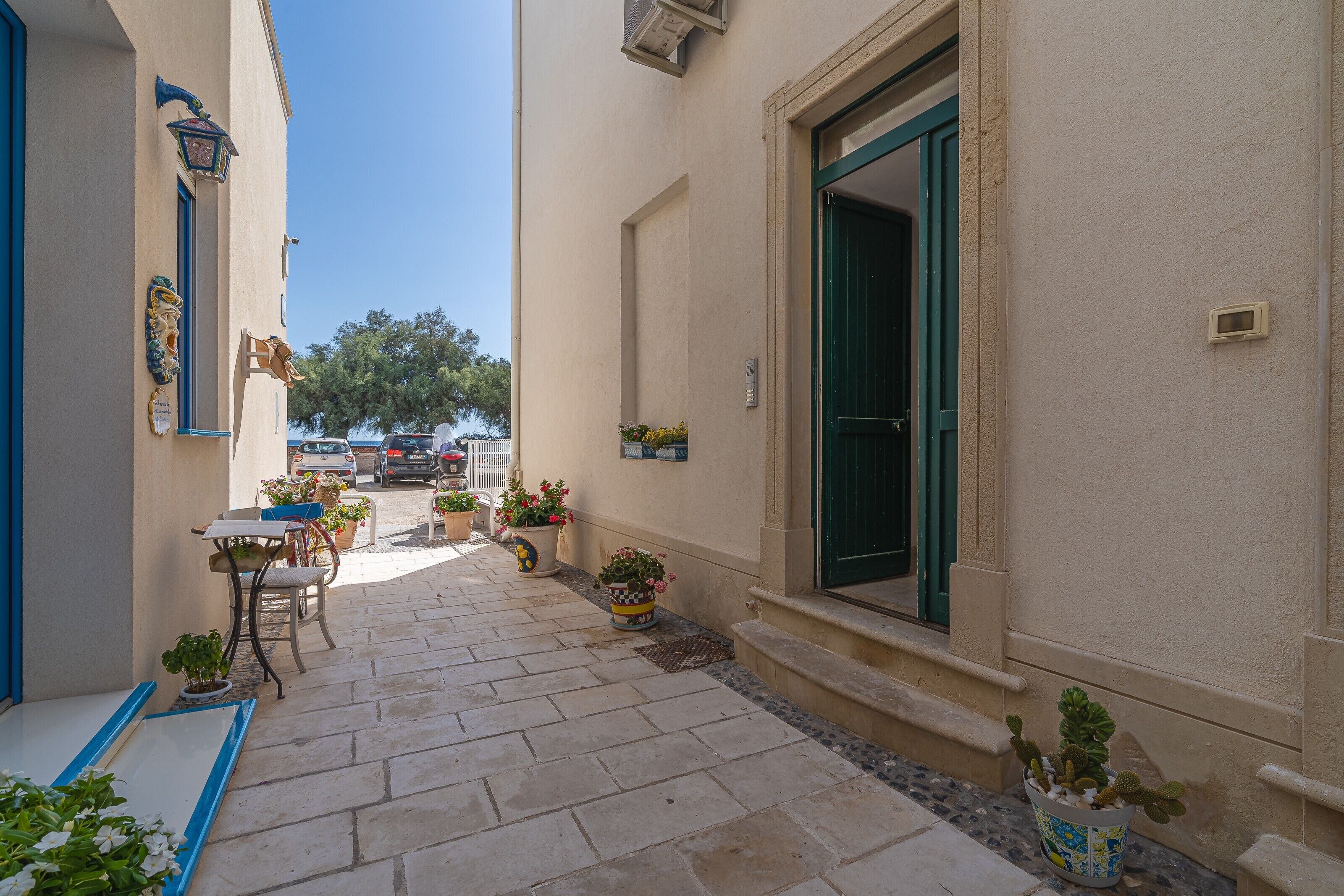 A narrow outdoor corridor with a table and chairs, potted plants, and a building entrance.