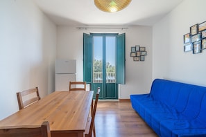 Una habitación con una mesa de comedor de madera, sillas, un sofá azul, una lámpara en el techo y una puerta con contraventanas verdes.