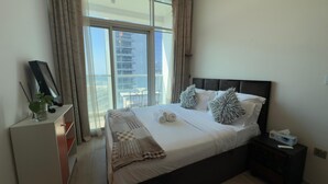 A hotel room with a large bed, a bedside table with a tissue box, a bedside lamp, and a view of a cityscape through large windows.