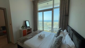 A hotel room with a large window offering a view of the sea and airport. There is a bed with white linens and a small bedside table with a mirror and a plant.