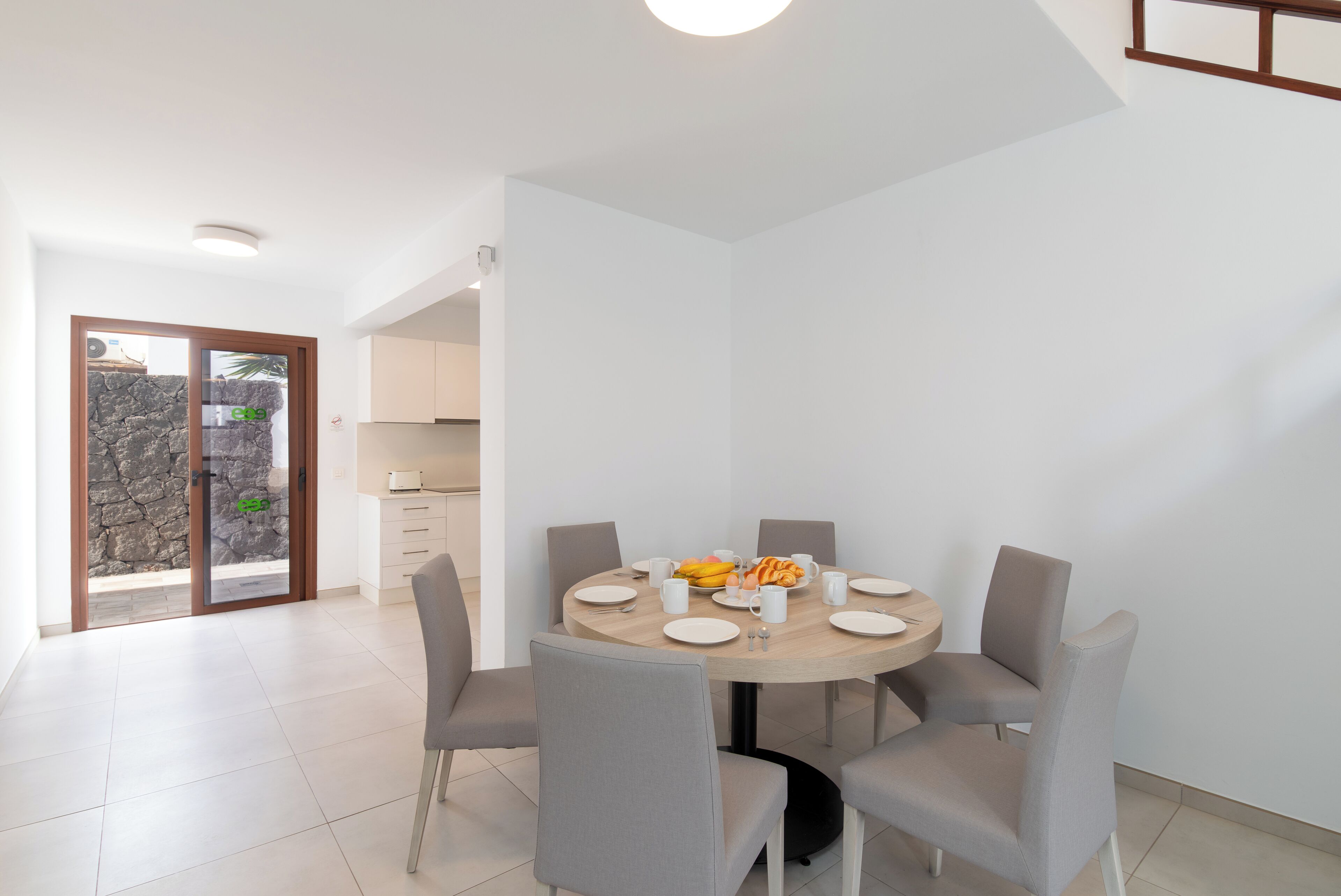 A modern dining area with a round table, grey chairs, and a kitchen in the background.