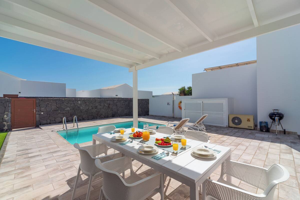 An outdoor dining area with a pool, white furniture, and a stone wall.