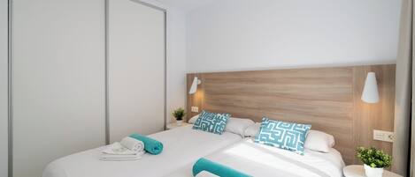 A hotel room with two beds, a wooden headboard, a small bedside table with a lamp, and a potted plant.
