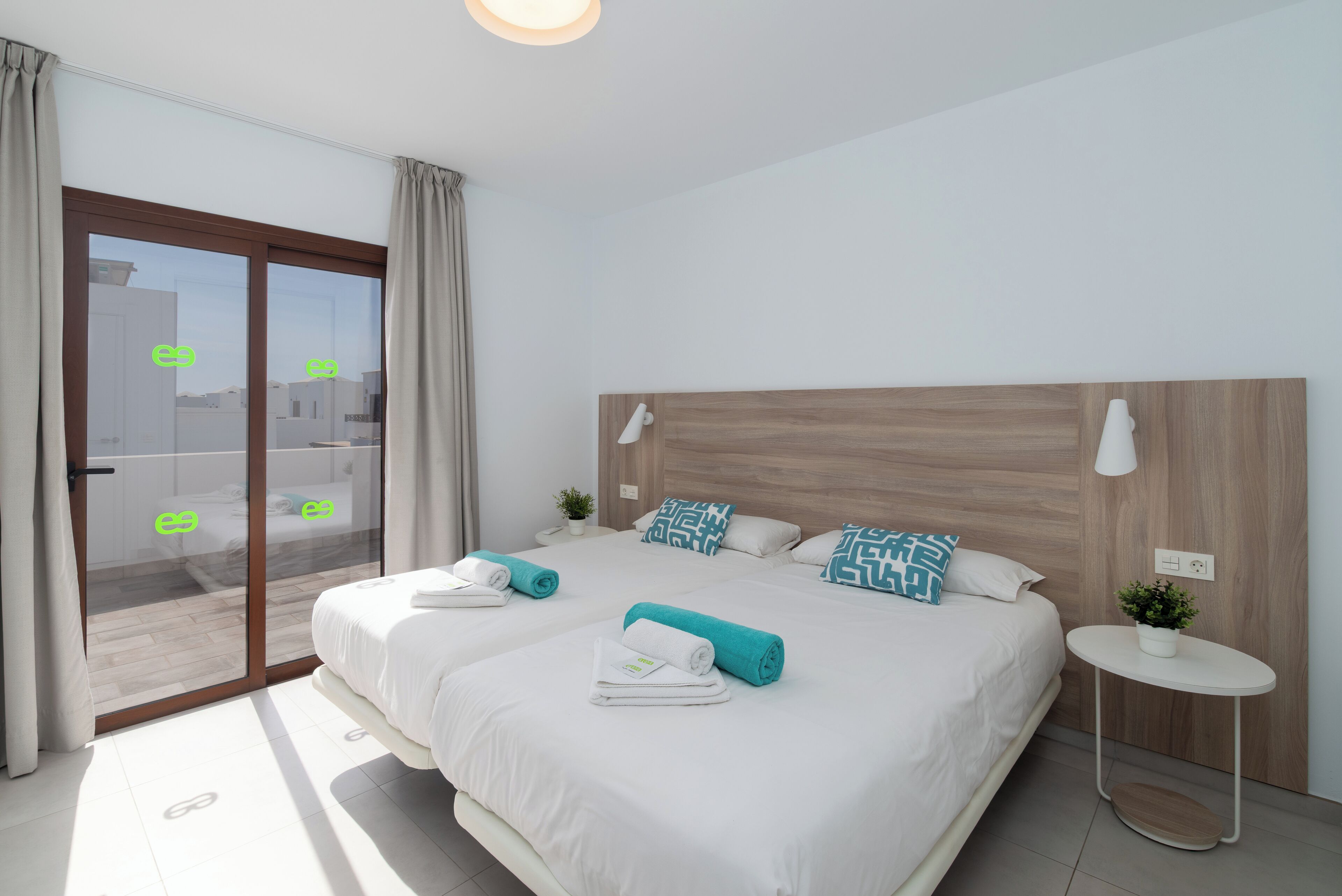 A modern hotel room with a large bed, two bedside tables, a small round table, and a sliding glass door leading to a balcony.