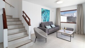 A modern living room with a staircase, a grey sofa, a coffee table, and a large window with curtains. - Ereza Villas La Granja (Yaiza)