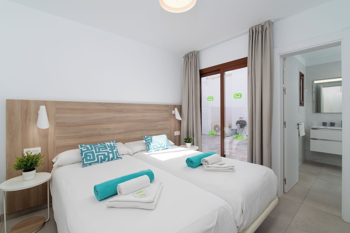 A modern hotel room with two beds, a small bedside table with a plant, a large window with curtains, and a bathroom visible through an open door.