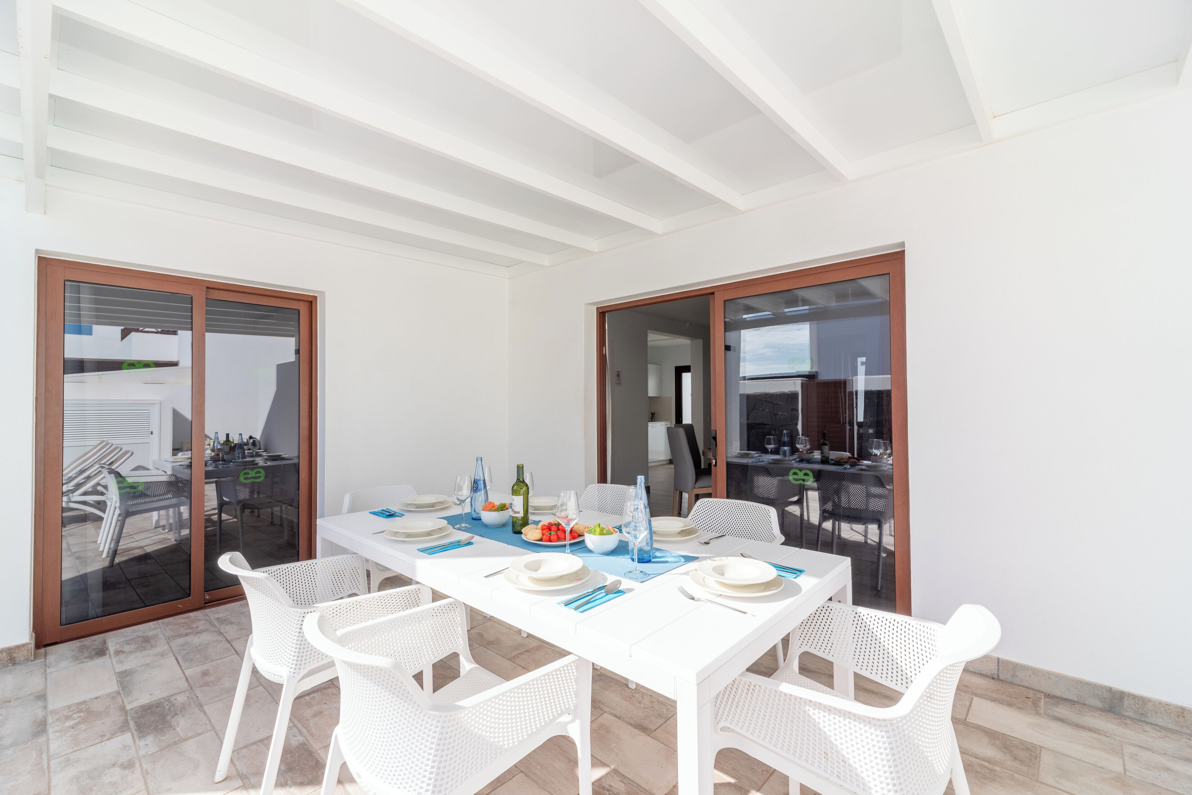 A dining area with a white table set for four, surrounded by white chairs, with a view of an outdoor patio through glass doors.