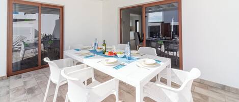 A dining area with a white table set for four, surrounded by white chairs, with a view of an outdoor patio through glass doors.