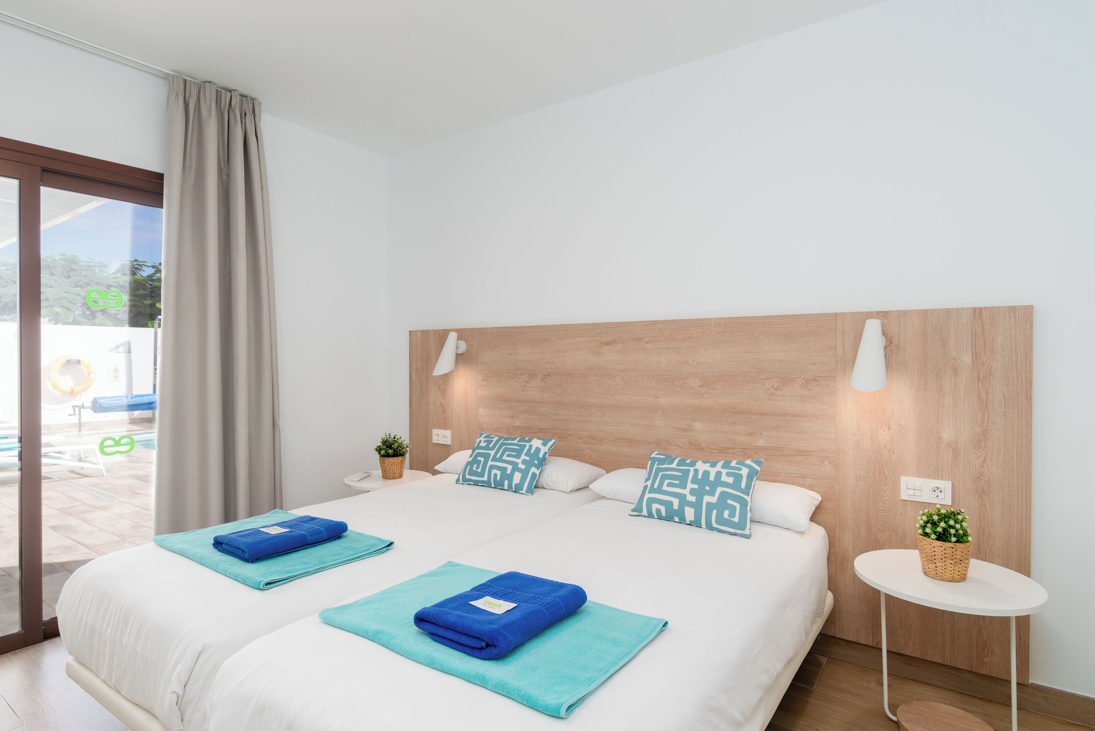 A modern hotel room with a large bed, two pillows with geometric patterns, a small bedside table with a plant, and a view of the outdoors through a sliding door.