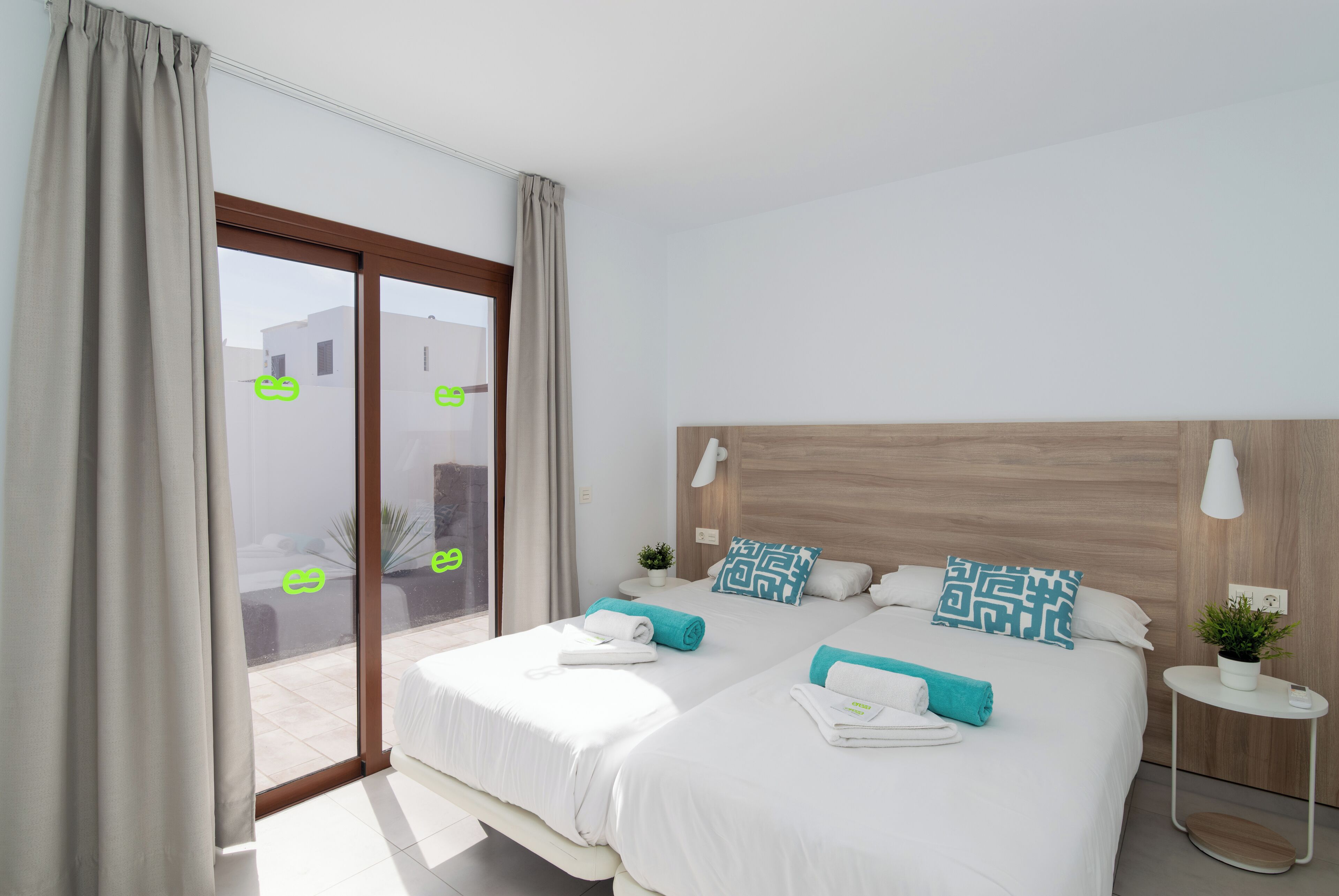 Habitación de hotel moderna con dos camas, un cabecero de madera, una mesita de noche pequeña con una lámpara y vistas a través de un gran ventanal corredero.