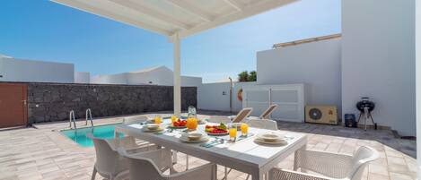 An outdoor patio with a pool, dining table, chairs, and a barbecue grill.