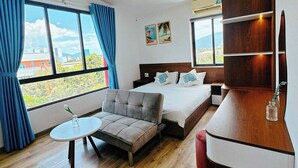 A hotel room with a bed, a sofa, a small table, and a wooden desk. There are large windows with curtains, a painting on the wall, and a view of buildings and mountains outside. - LOUIS OTIS APARTMENT 03 (Da Nang)