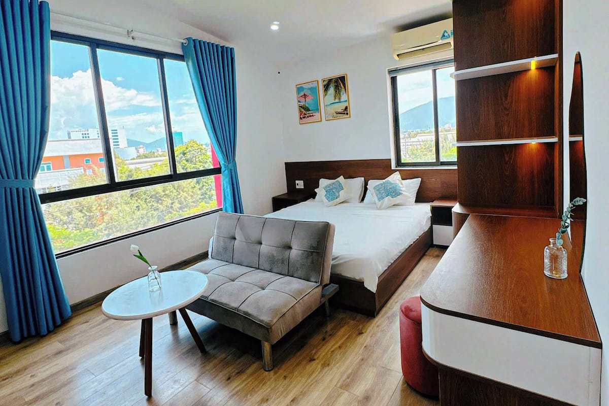Une chambre d’hôtel avec un lit, un canapé, une petite table et un bureau en bois. De grandes fenêtres sont présentes, avec des rideaux, ainsi qu’un tableau au mur. La vue donne sur des bâtiments et des montagnes.