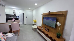 A modern living room with a flat-screen TV mounted on a wooden wall, a dining area with pink chairs, and a kitchen in the background. - Flat Centro Porto de Galinhas 204 byLP (Ipojuca)