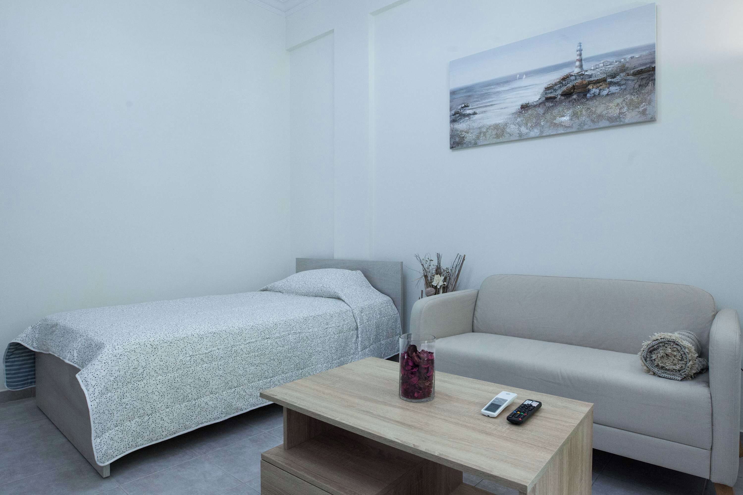 Una habitación pequeña y moderna con una cama, un sofá y una mesita de centro. Hay un cuadro en la pared y un jarrón sobre la mesa.