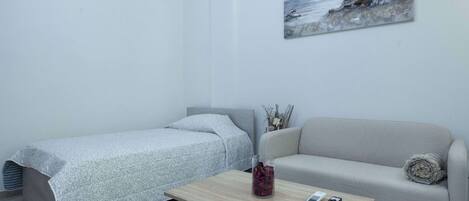 Una habitación pequeña y moderna con una cama, un sofá y una mesita de centro. Hay un cuadro en la pared y un jarrón sobre la mesa.
