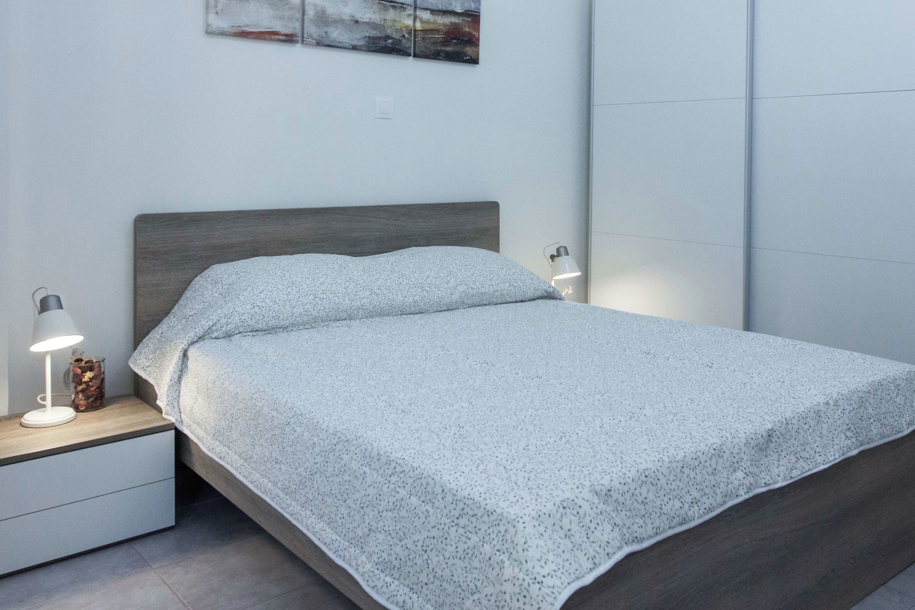 Una cama bien hecha con una colcha gris estampada y almohadas, flanqueada por mesitas de noche con lámparas y un pequeño objeto decorativo.