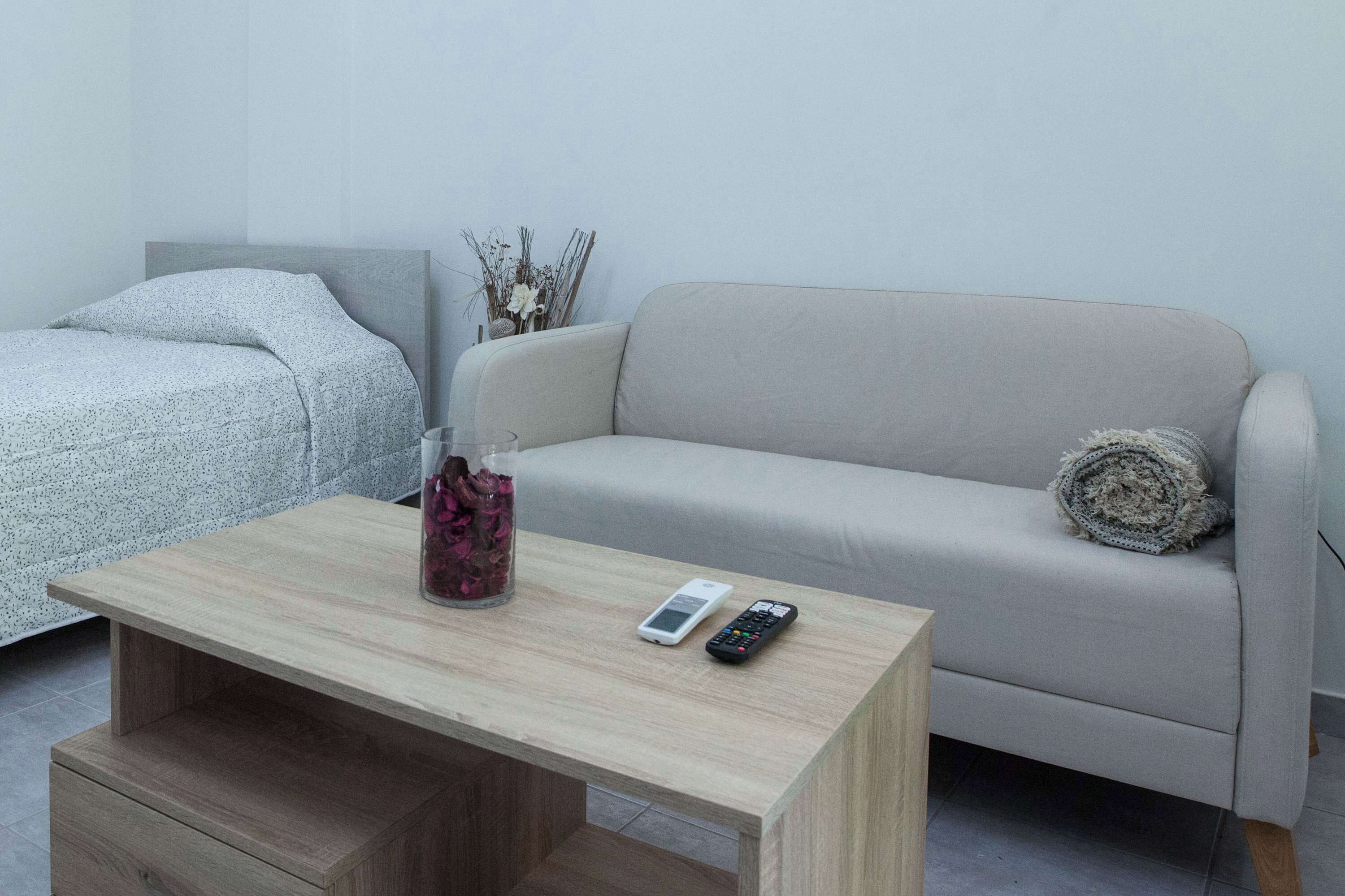 A minimalist living room with a light-colored sofa, a wooden coffee table, and a bed with a patterned cover.