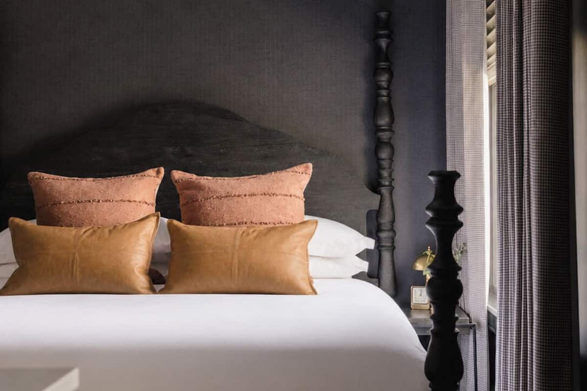 Una cama bien hecha con dos almohadas, cabecera oscura y mesita de noche con lámpara.