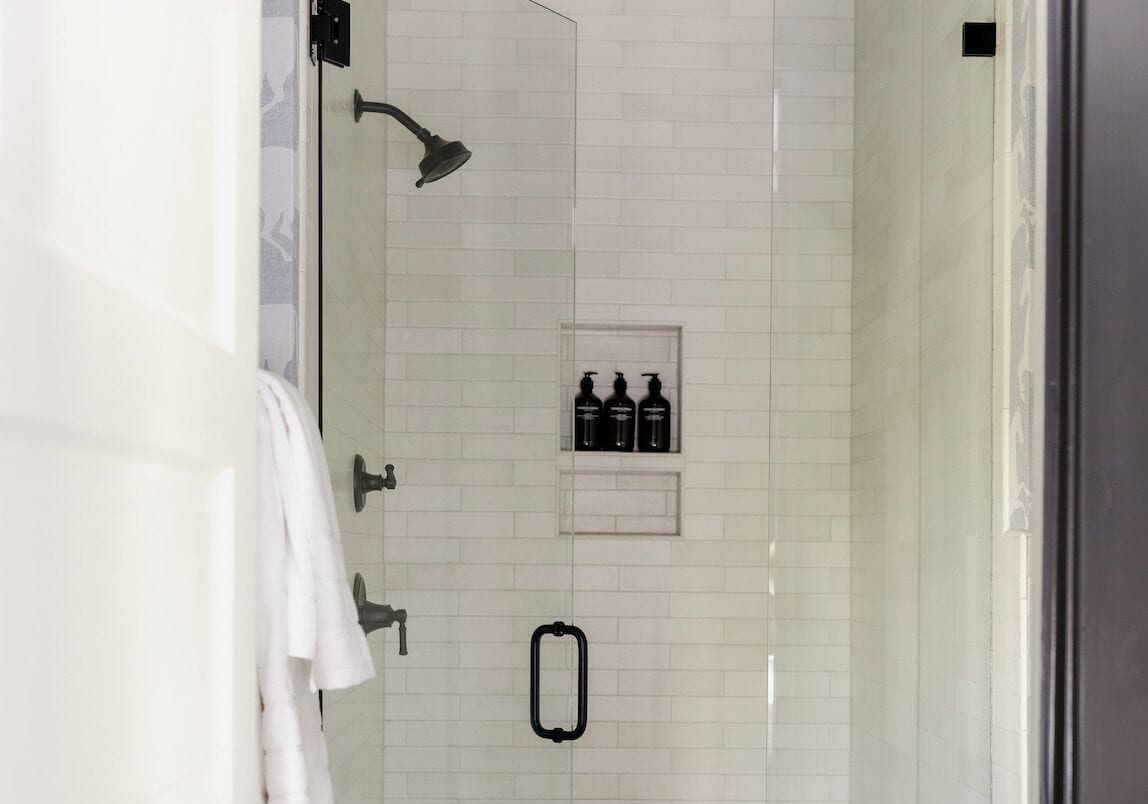 A modern bathroom with white tiles, a shower, a towel rack, and a shelf with toiletries.