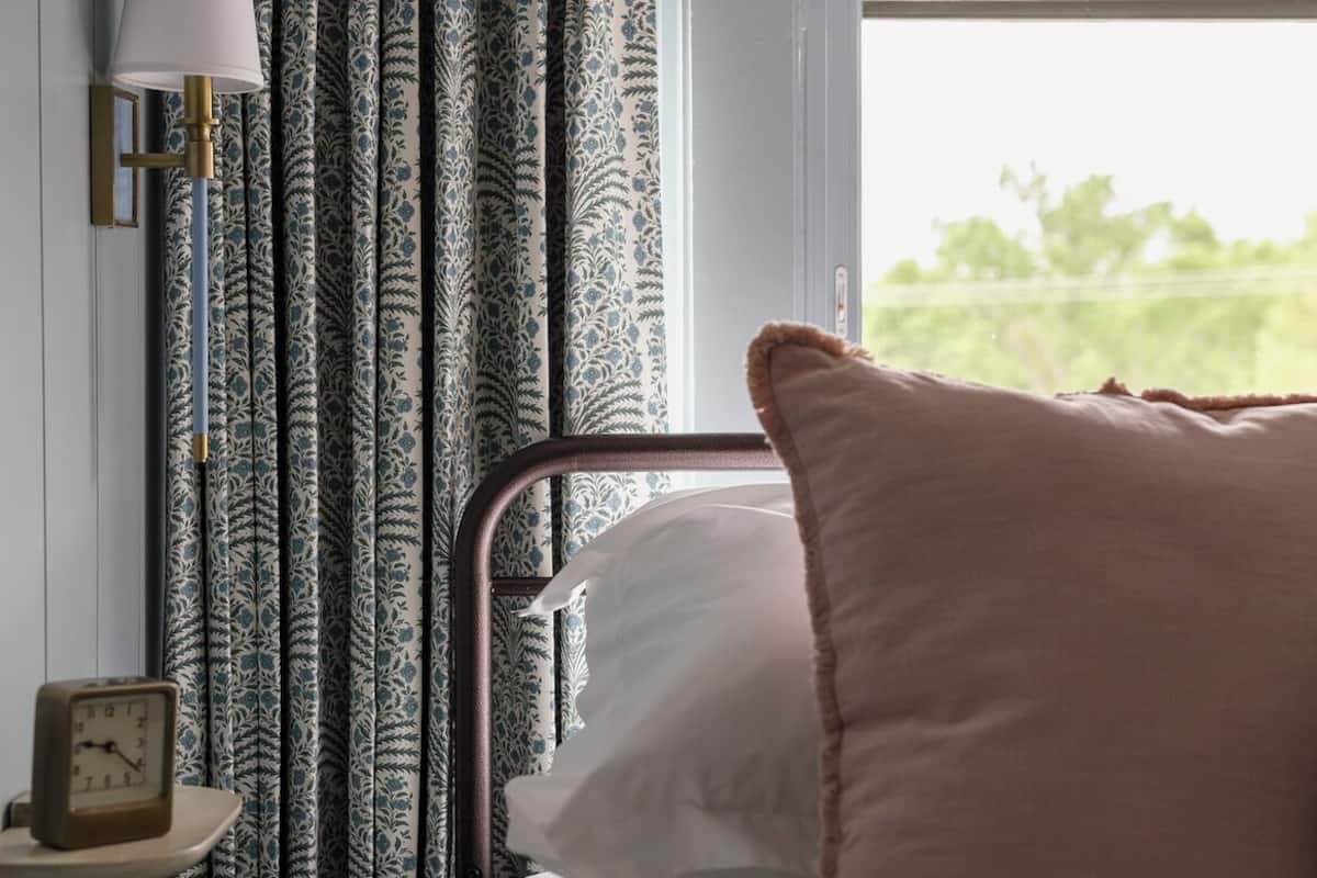 Una cama con un cojín blanco, una lámpara de noche y cortinas con estampado de hojas.