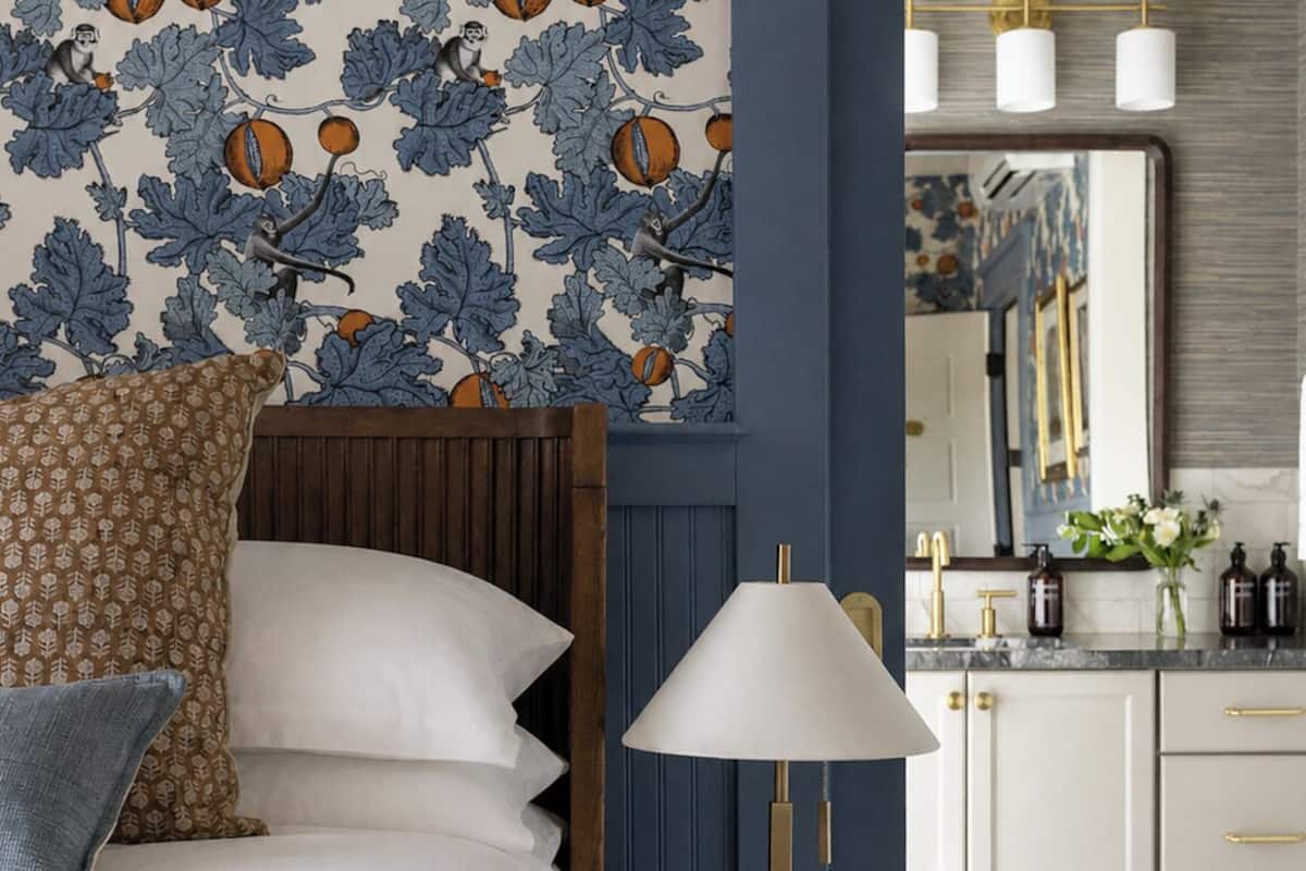 Un dormitorio con cama, almohadas, una mesita de noche con lámpara, un reloj, y un baño con espejo, lavabo y armarios.