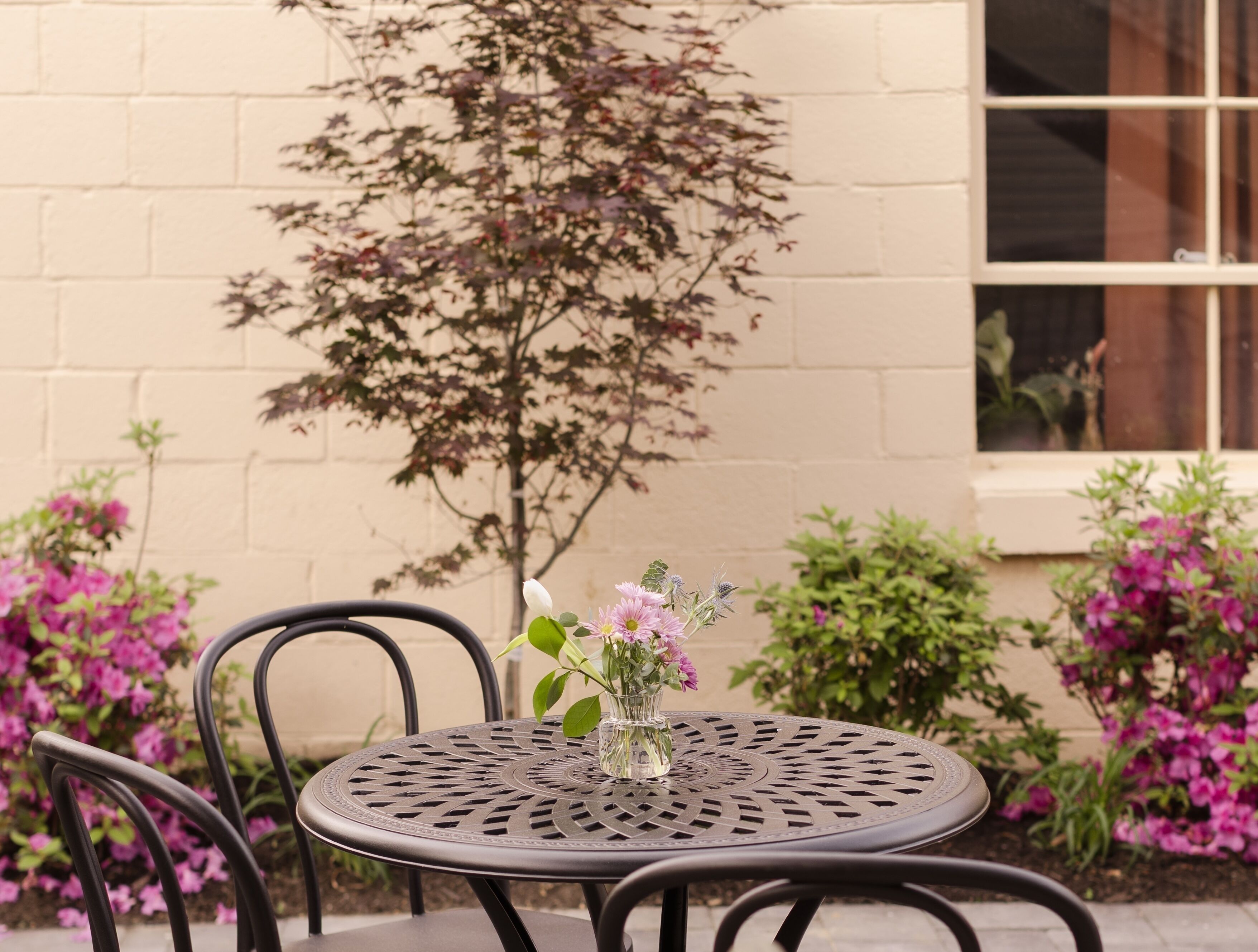 A round metal table with a glass vase of flowers, surrounded by chairs, against a beige wall with a window and a tree.