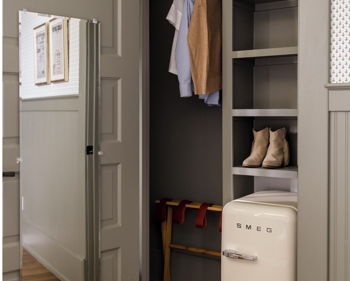 A room with a wardrobe, a coat rack with clothes, a pair of boots, and a white appliance with 'SMEG' on it.