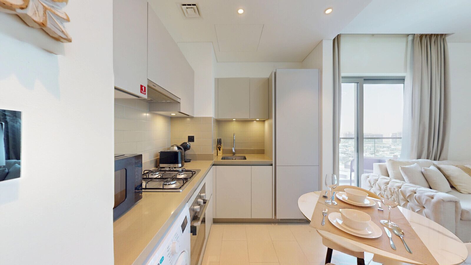 A modern kitchen with a gas stove, a microwave, and a sink. There is a dining area with a table set for two, and a balcony with a view.