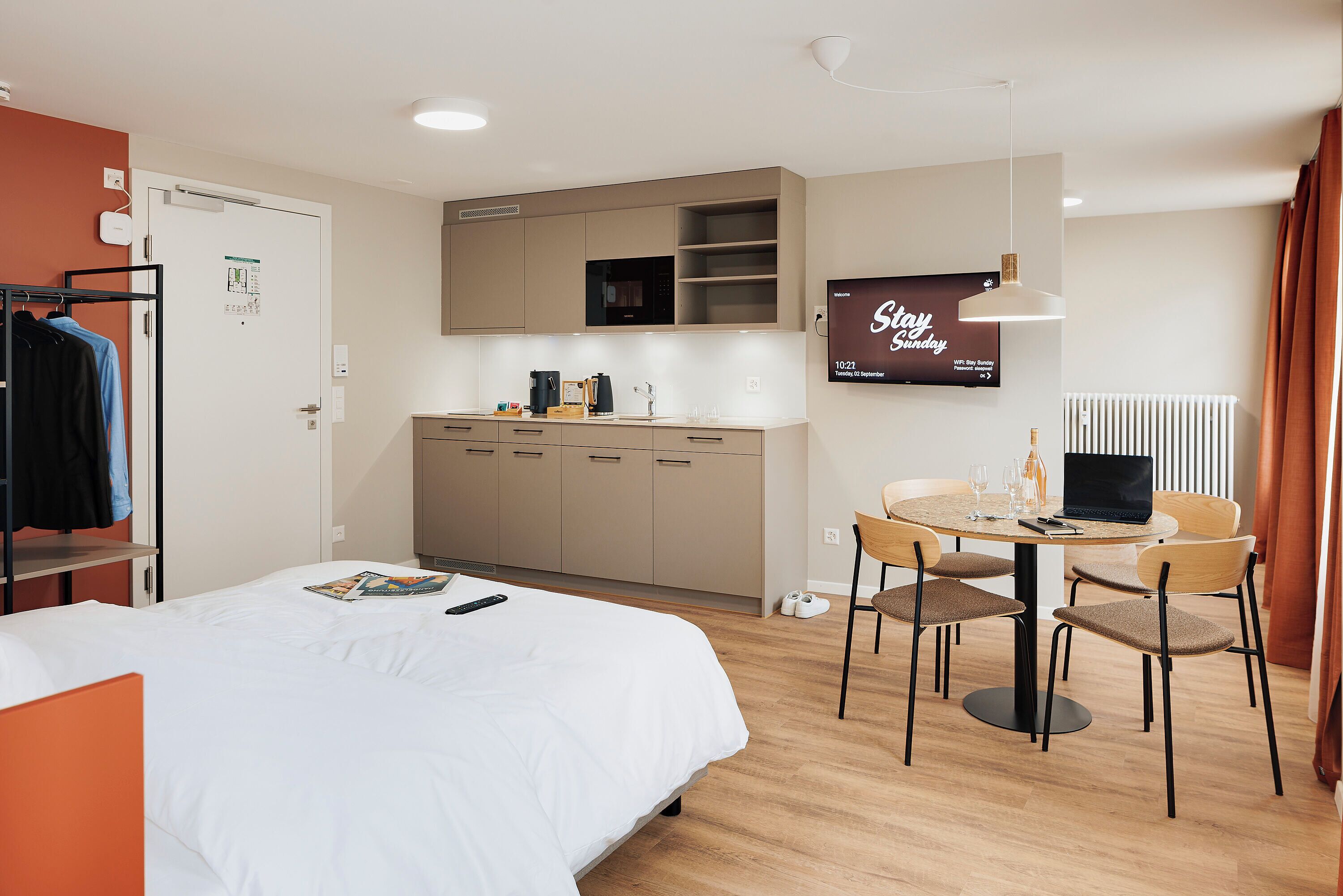 Ein Hotelzimmer mit einem Bett, einem Schreibtisch mit einem Laptop, einem Fernseher, der 'Stay Sunday' anzeigt, und einer Küchenzeile.