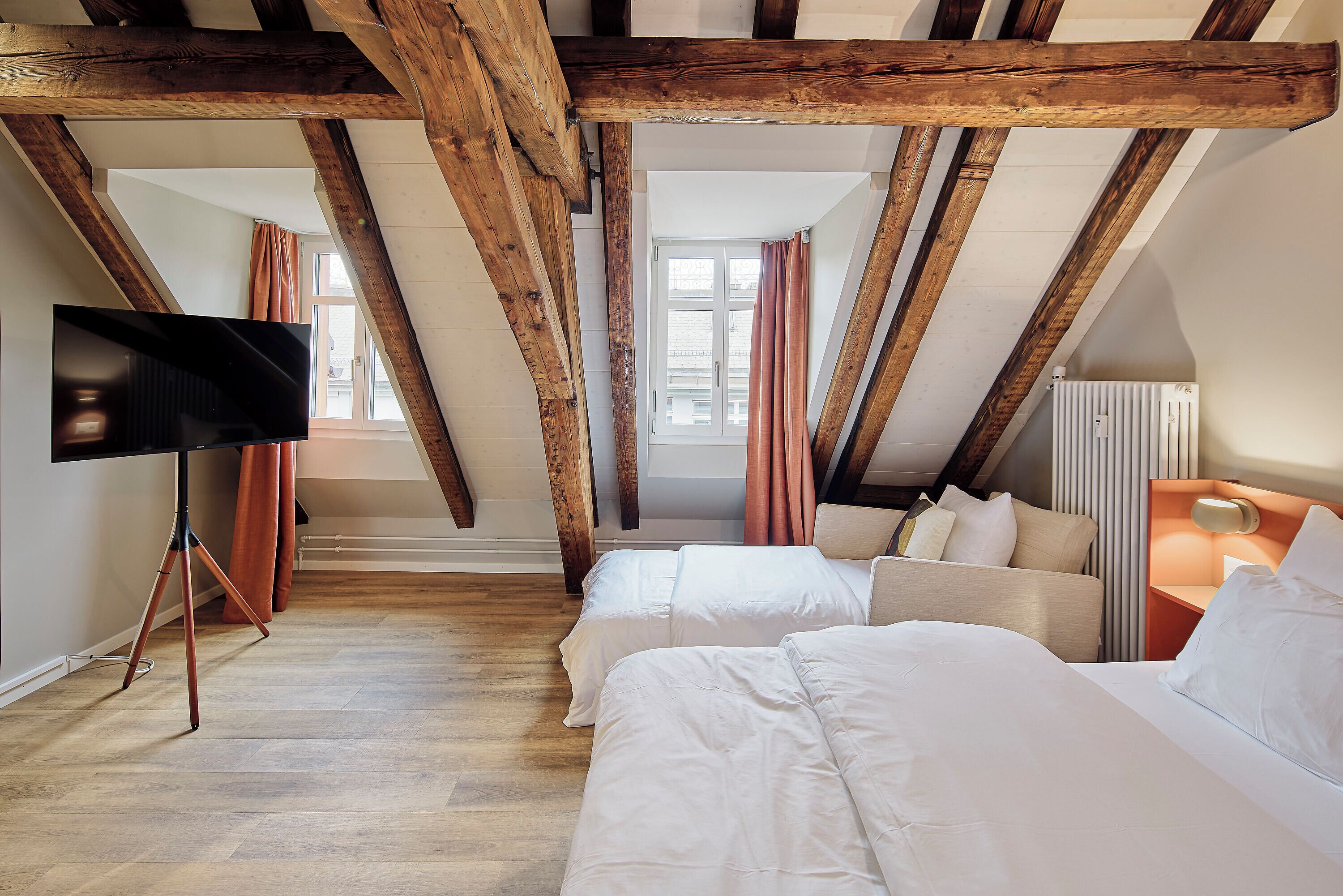 A hotel room with exposed wooden beams, two beds, a television on a tripod stand, and a window with curtains.