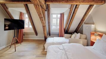 Ein Hotelzimmer mit sichtbaren Holzbalken, zwei Betten, einem Fernseher auf einem Stativ und einem Fenster mit Vorhängen.