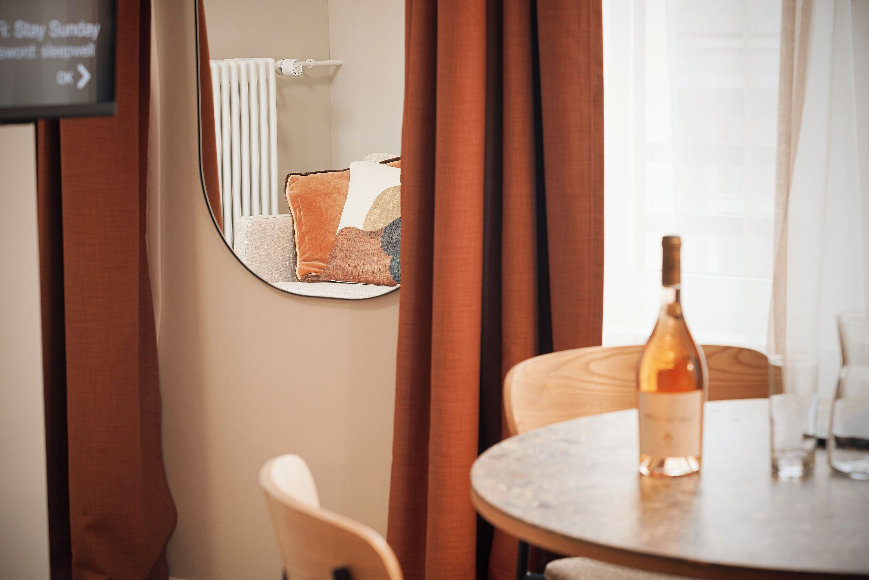 Habitación de hotel con una mesa redonda, una botella de vino y un espejo que refleja una cama con almohadillas.